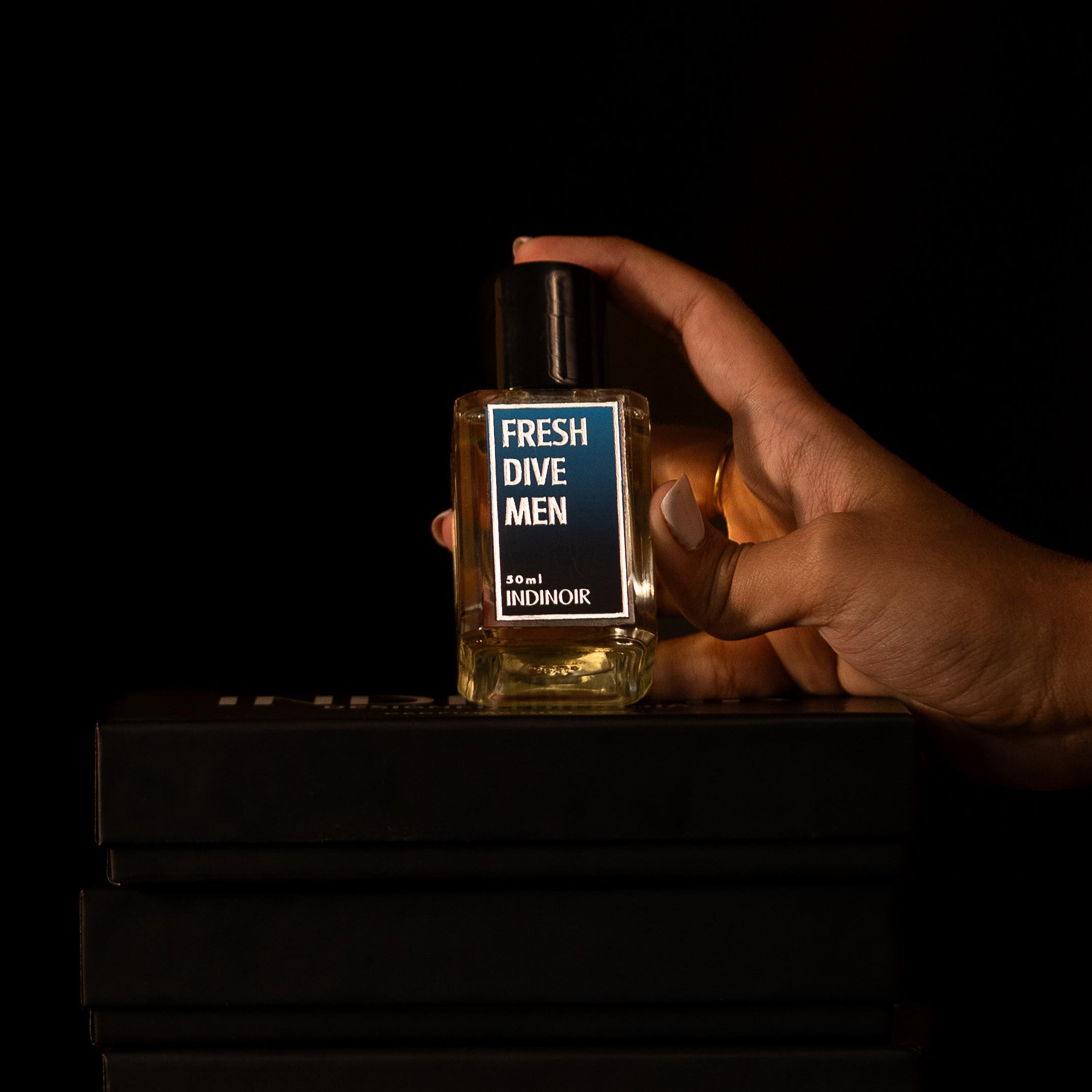 Hand holding a bottle of 'Fresh Dive Men' cologne on black boxes with a dark background