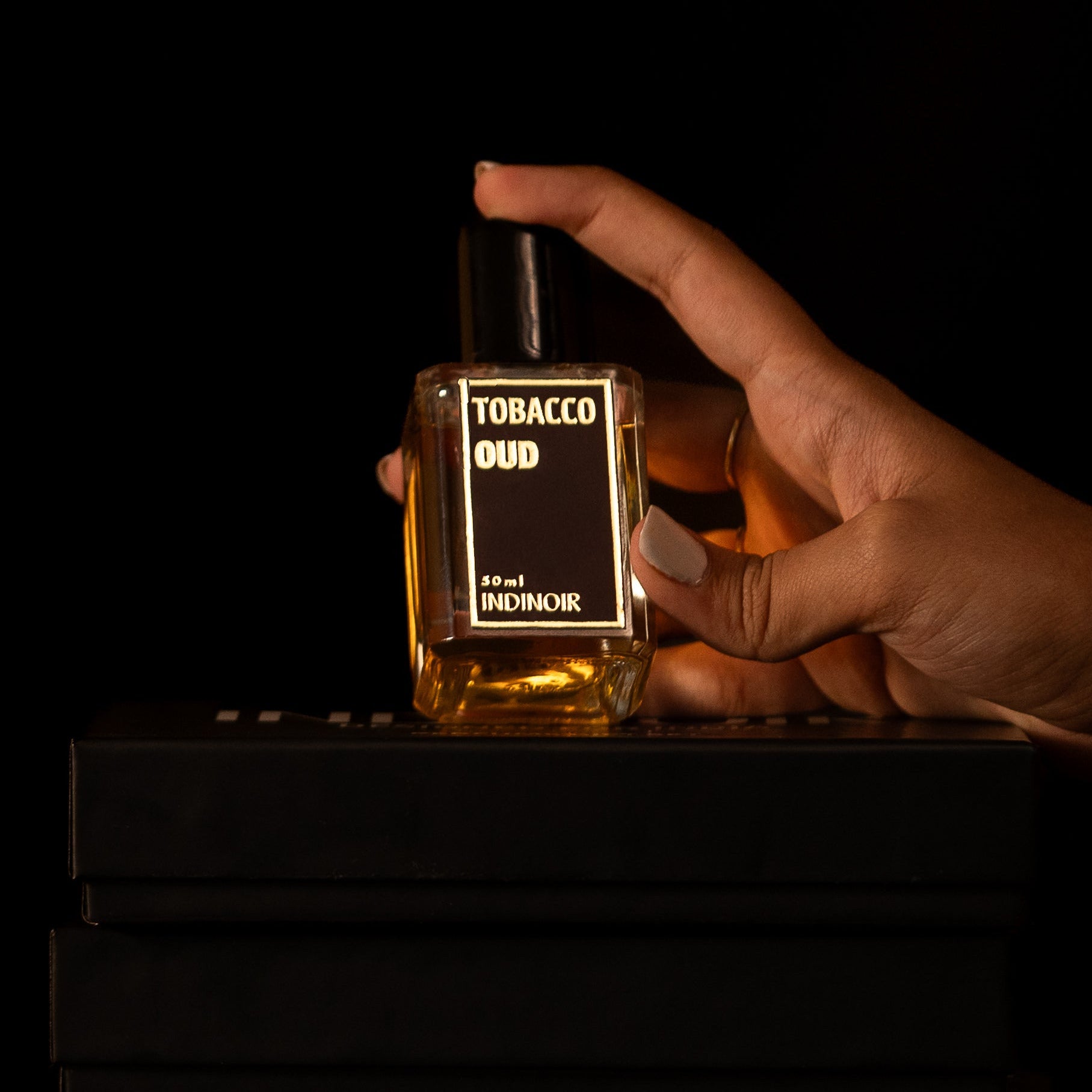 Hand holding a perfume bottle labeled 'Tobacco Oud' against a dark background